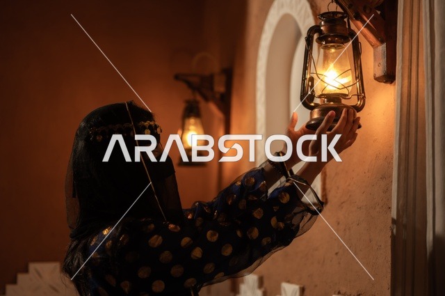 Expressing feelings of love for the homeland and belonging to it, pride in national identity, a Gulf Arab Saudi woman wearing a black abaya and a niqab holding a metal lantern in an old heritage house, on the day we began in 1727 AD, the anniversary of the founding of the first Saudi state on February 22, wearing traditional folk costumes.