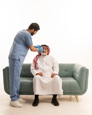 شركات التمريض المنزلي, الحاجة إلى المساعدة الطبية, بورتريه لرجل مسن عربي خليجي سعودي يرتدي الثوب التقليدي والشماغ يجلس على الكنبة مصاب بالحمى, ممرض يلبس الكفوف وكمامة الوقاية يقوم بقياس درجة حرارة الجسم بواسطة جهاز الترمومتر الرقمي، خلفية بيضاء