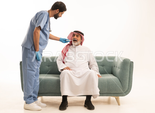 Home nursing companies, need for medical assistance, portrait of an elderly Saudi Arabian Gulf man wearing traditional thobe and shemagh sitting on a sofa with a fever, a nurse wearing gloves and a protective mask is measuring body temperature with a digital thermometer, white background