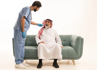 Home nursing companies, need for medical assistance, portrait of an elderly Saudi Arabian Gulf man wearing traditional thobe and shemagh sitting on a sofa with a fever, a nurse wearing gloves and a protective mask is measuring body temperature with a digital thermometer, white background