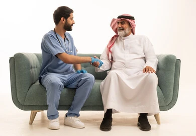 تقديم أفضل الخدمات الصحية لمرضى كبار السن في المستشفى، بورتريه لممرض يرتدي اليونيفورم الخاص والقفازات والكمامة الوقائية يقوم بطمأنة رجل عربي خليجي سعودي يرتدي الزي التقليدي والشماغ، مفهوم الطب والرعاية الصحية، تقدير الطبيب لجهوده ورعايته الطبية، خلفية رمادية