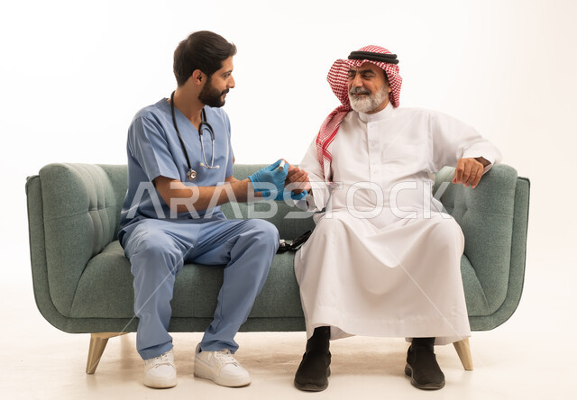 جهاز قياس سكر الدم لمرضى السكري، العمل في القطاع الطبي وشركات التمريض المنزلي، الرعاية الصحية والفحص الطبي السريع، بورتريه لممرض يرتدي المعطف الطبي يقوم بفحص نسبة السكر في الدم لمريض مسن عربي خليجي سعودي يرتدي الشماغ والثوب التقليدي يجلس على الكنبه، خلفية بيضاء