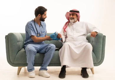 Blood glucose meter for diabetics, working in the medical sector and home nursing companies, healthcare and rapid medical examination, portrait of a nurse wearing a medical coat checking the blood sugar level of an elderly Saudi Arabian Gulf Arab patient wearing a shemagh and traditional thobe sitting on a sofa, white background