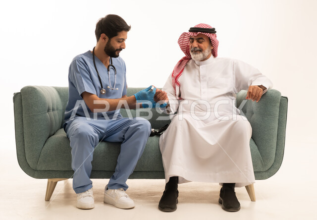 جهاز قياس سكر الدم لمرضى السكري، العمل في القطاع الطبي وشركات التمريض المنزلي، الرعاية الصحية والفحص الطبي السريع، بورتريه لممرض يرتدي المعطف الطبي يقوم بفحص نسبة السكر في الدم لمريض مسن عربي خليجي سعودي يرتدي الشماغ والثوب التقليدي يجلس على الكنبه، خلفية بيضاء
