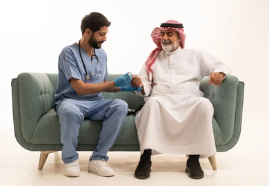 Blood glucose meter for diabetics, working in the medical sector and home nursing companies, healthcare and rapid medical examination, portrait of a nurse wearing a medical coat checking the blood sugar level of an elderly Saudi Arabian Gulf Arab patient wearing a shemagh and traditional thobe sitting on a sofa, white background