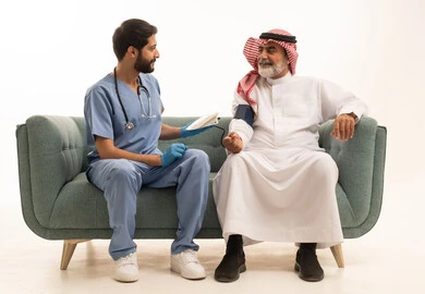 الخدمات الطبية والرعاية الصحية، بورتريه لممرض يرتدي اليونيفورم والقفازات والكمامة الوقائية يقوم بقياس ضغط الدم لرجل مسن عربي خليجي سعودي يلبس الثوب التقليدي والشماغ يجلس على كرسي مريح، الإصابة بوعكة صحية، العمل في القطاع الصحي، خلفية بيضاء