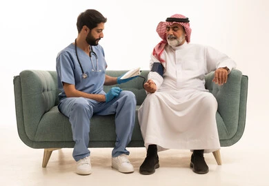 الخدمات الطبية والرعاية الصحية، بورتريه لممرض يرتدي اليونيفورم والقفازات والكمامة الوقائية يقوم بقياس ضغط الدم لرجل مسن عربي خليجي سعودي يلبس الثوب التقليدي والشماغ يجلس على كرسي مريح، الإصابة بوعكة صحية، العمل في القطاع الصحي، خلفية بيضاء