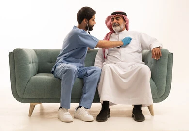استخدام سماعة الطبيب لسماع نبضات القلب، مفهوم الطب والرعاية الصحية، بورتريه لممرض يرتدي اليونيفورم والقفازات والكمامة الوقائية يقوم بفحص رجل مسن عربي خليجي سعودي يلبس الثوب التقليدي والشماغ يجلس على كرسي مريح، تقديم الخدمات الصحية للمرضى، خلفية بيضاء