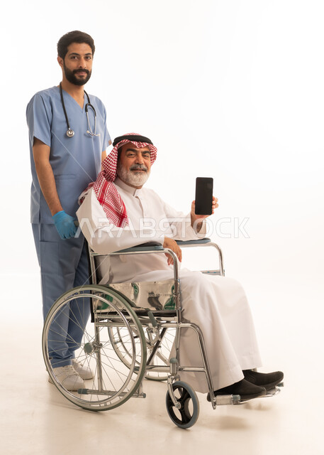 عرض شاشة هاتف محمول سوداء فارغة, مفهوم الرعاية الصحية والاعتناء بالمرضى والمقعدين، نمط حياة ذوي الهمم، بورتريه لممرض يرتدي اليونيفورم والكمامة يقوم بالاعتناء برجل عربي خليجي سعودي مقعد يرتدي الثوب التقليدي والشماغ يجلس على كرسي متحرك، صورة لكامل الجسم، خلفية بيضاء