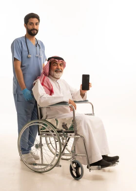 عرض شاشة هاتف محمول سوداء فارغة, مفهوم الرعاية الصحية والاعتناء بالمرضى والمقعدين، نمط حياة ذوي الهمم، بورتريه لممرض يرتدي اليونيفورم والكمامة يقوم بالاعتناء برجل عربي خليجي سعودي مقعد يرتدي الثوب التقليدي والشماغ يجلس على كرسي متحرك، صورة لكامل الجسم، خلفية بيضاء
