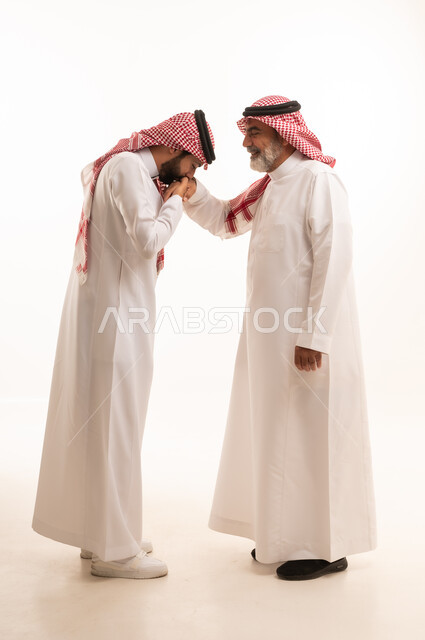 Expressions of appreciation and respect for elders, adherence to customs and traditions, the concept of family ties and kinship, a refined family atmosphere, a portrait of a young Saudi Arabian Gulf man wearing traditional thobe and shemagh kissing his father's hand, gestures of greeting and peace, full body, white background