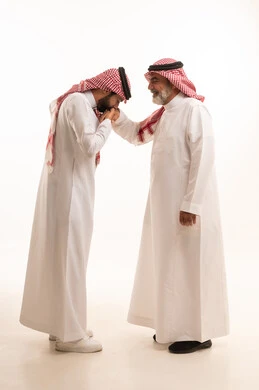 Expressions of appreciation and respect for elders, adherence to customs and traditions, the concept of family ties and kinship, a refined family atmosphere, a portrait of a young Saudi Arabian Gulf man wearing traditional thobe and shemagh kissing his father's hand, gestures of greeting and peace, full body, white background