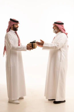 تقديم الهدايا التذكارية للآباء، التعبير عن المحبة والألفة في المناسبات، بورتريه لشاب عربي خليجي سعودي يرتدي الثوب التقليدي والشماغ يقوم بتقديم هدية لوالده، ايماءات الفرح والسعادة والمحبة، أعياد واحتفالات وأجواء سعيدة، الجسم بالطول الكامل, خلفية بيضاء