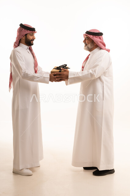 تقديم الهدايا التذكارية للآباء، التعبير عن المحبة والألفة في المناسبات، بورتريه لشاب عربي خليجي سعودي يرتدي الثوب التقليدي والشماغ يقوم بتقديم هدية لوالده، ايماءات الفرح والسعادة والمحبة، أعياد واحتفالات وأجواء سعيدة، الجسم بالطول الكامل, خلفية بيضاء