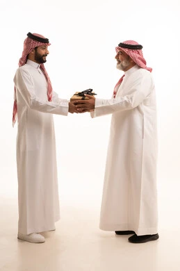 تقديم الهدايا التذكارية للآباء، التعبير عن المحبة والألفة في المناسبات، بورتريه لشاب عربي خليجي سعودي يرتدي الثوب التقليدي والشماغ يقوم بتقديم هدية لوالده، ايماءات الفرح والسعادة والمحبة، أعياد واحتفالات وأجواء سعيدة، الجسم بالطول الكامل, خلفية بيضاء