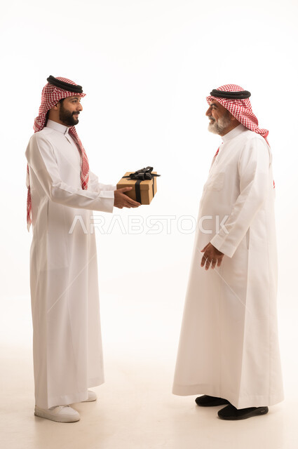 تقديم الهدايا التذكارية للآباء، التعبير عن المحبة والألفة في المناسبات، بورتريه لشاب عربي خليجي سعودي يرتدي الثوب التقليدي والشماغ يقوم بتقديم هدية لوالده، ايماءات الفرح والسعادة والمحبة، أعياد واحتفالات وأجواء سعيدة، الجسم بالطول الكامل, خلفية بيضاء