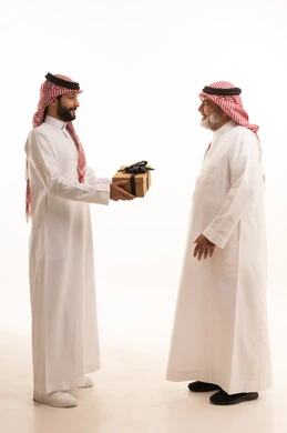 تقديم الهدايا التذكارية للآباء، التعبير عن المحبة والألفة في المناسبات، بورتريه لشاب عربي خليجي سعودي يرتدي الثوب التقليدي والشماغ يقوم بتقديم هدية لوالده، ايماءات الفرح والسعادة والمحبة، أعياد واحتفالات وأجواء سعيدة، الجسم بالطول الكامل, خلفية بيضاء