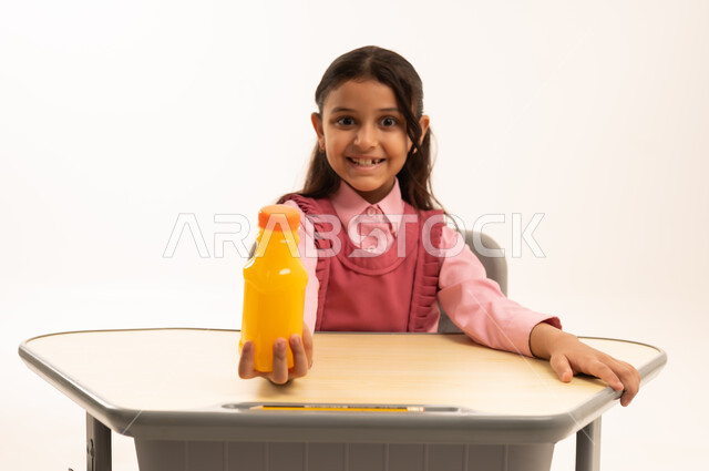 شرب العصير الصحي في الفسحة المدرسية، الاستمتاع في وقت الفسحة المدرسية، موسم العودة للمدارس، بورتريه لطالبة عربيه خليجيه سعودية ترتدي الزي المدرسة تجلس على الطاوله المكتبية تمسك بيدها زجاجة عصير برتقال طبيعي، خلفية بيضاء