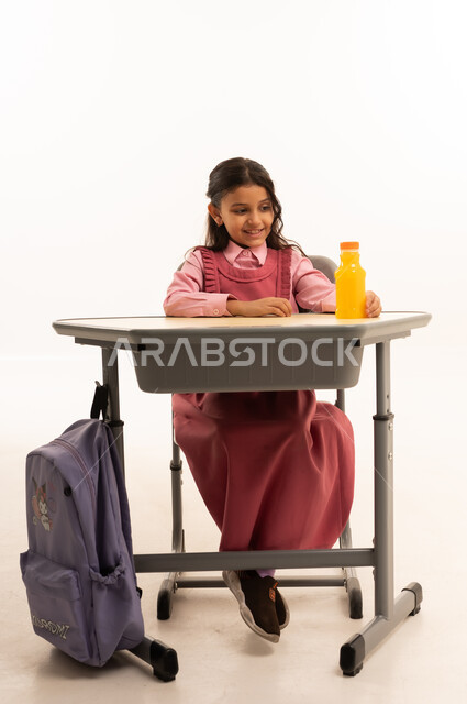 شرب العصير الصحي في الفسحة المدرسية، الاستمتاع في وقت الفسحة المدرسية، موسم العودة للمدارس، بورتريه لطالبة عربيه خليجيه سعودية ترتدي الزي المدرسة تجلس على الطاوله المكتبية تمسك بيدها زجاجة عصير برتقال طبيعي، خلفية بيضاء