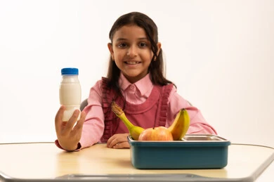 الاستمتاع بتناول الأطعمة الصحية في وقت الفسحة المدرسية، موسم العودة للمدارس، بورتريه مقرب لطالبة عربيه خليجيه سعودية ترتدي الزي المدرسة تجلس على الطاوله المكتبية، تناول وجبة الفطور الصحية في الفسحة المدرسية، خلفية بيضاء