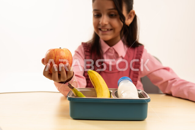 hahdvEnjoying healthy food during school break, back to school season, close-up portrait of a Saudi Arabian Gulf Arab female student wearing school uniform sitting at a desk, eating a healthy breakfast during school break, white background