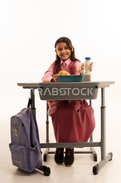  تناول وجبة الفطور الصحية في الفسحة المدرسية، الاستمتاع بتناول الأطعمة الصحية في وقت الفسحة المدرسية، موسم العودة للمدارس، بورتريه لطالبة عربيه خليجيه سعودية ترتدي الزي المدرسة تجلس على الطاوله المكتبية، خلفية بيضاء
