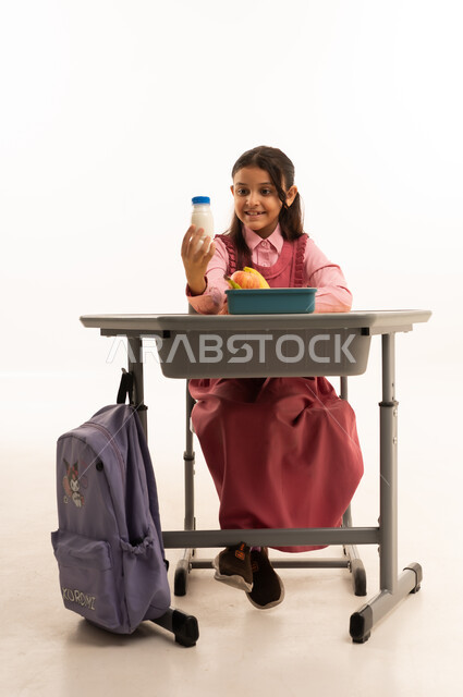  تناول وجبة الفطور الصحية في الفسحة المدرسية، الاستمتاع بتناول الأطعمة الصحية في وقت الفسحة المدرسية، موسم العودة للمدارس، بورتريه لطالبة عربيه خليجيه سعودية ترتدي الزي المدرسة تجلس على الطاوله المكتبية، خلفية بيضاء