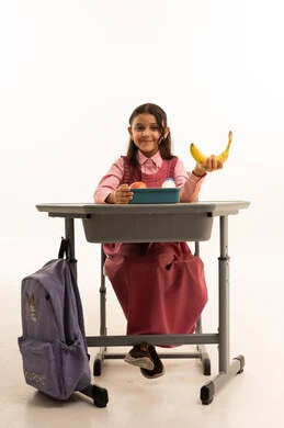  تناول وجبة الفطور الصحية في الفسحة المدرسية، الاستمتاع بتناول الأطعمة الصحية في وقت الفسحة المدرسية، موسم العودة للمدارس، بورتريه لطالبة عربيه خليجيه سعودية ترتدي الزي المدرسة تجلس على الطاوله المكتبية، خلفية بيضاء