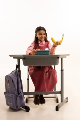  تناول وجبة الفطور الصحية في الفسحة المدرسية، الاستمتاع بتناول الأطعمة الصحية في وقت الفسحة المدرسية، موسم العودة للمدارس، بورتريه لطالبة عربيه خليجيه سعودية ترتدي الزي المدرسة تجلس على الطاوله المكتبية، خلفية بيضاء