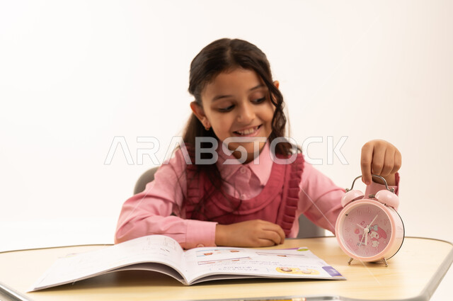 مفهوم تنظيم و إدارة الوقت, الاستعداد للعام الدراسي الجديد, التعليم في المملكة العربية السعودية, بورتريه لفتاة عربية خليجية سعودية ترتدي الزي المدرسي  تمسك بيدها ساعة المنبه تجلس على طاولتها المكتبية, خلفية بيضاء