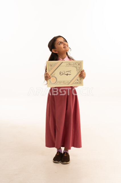 النجاح وانتهاء العام الدراسي، مفهوم التفوق والتخرج من المدرسة, النظر الى شيء ما بايماءات الفخر والسعاده, جودة التعليم في السعوديه, بورتريه  لطالبة عربية سعودية خليجية مبتسمة ترتدي الزي المدرسي تمسك بيدها الشهادة، الجسم بالطول الكامل، خلفية بيضاء