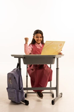 انتهاء العام الدراسي، مفهوم التفوق والنجاح والتخرج من المدرسة, النظر الى الكاميرا بايماءات الفخر والسعاده, جودة التعليم في السعوديه, بورتريه  لطالبة عربية سعودية خليجية مبتسمة ترتدي الزي المدرسي تجلس على مكتبها تمسك بيدها الشهادة، خلفية بيضاء