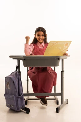 انتهاء العام الدراسي، مفهوم التفوق والنجاح والتخرج من المدرسة, النظر الى الكاميرا بايماءات الفخر والسعاده, جودة التعليم في السعوديه, بورتريه  لطالبة عربية سعودية خليجية مبتسمة ترتدي الزي المدرسي تجلس على مكتبها تمسك بيدها الشهادة، خلفية بيضاء