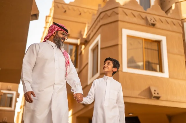 Enjoying playing with the family, a family atmosphere filled with affection, a family standing at the entrance of a modern home, a child wearing a white dress, having a good time with the children, sharing happy moments and fun with the children, an Arab Gulf Saudi father holding his son's hand and playing with him, the concept of a happy family.