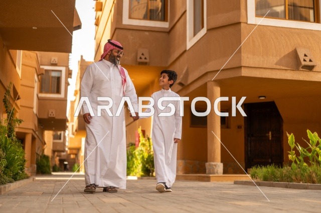 Enjoying playing with the family, a family atmosphere filled with affection, a family standing at the entrance of a modern home, a child wearing a white dress, having a good time with the children, sharing happy moments and fun with the children, an Arab Gulf Saudi father holding his son's hand and playing with him, the concept of a happy family.