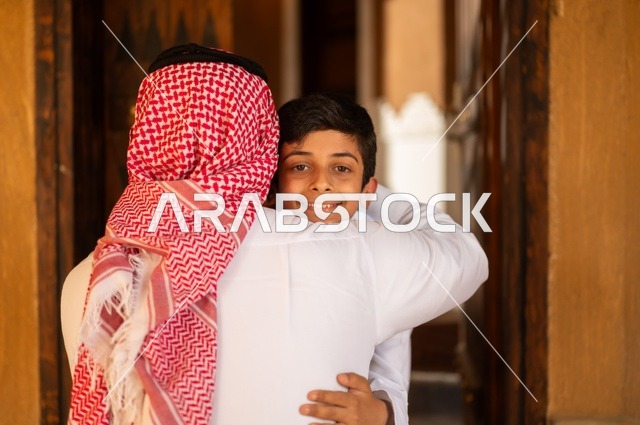 العناق والاستقبال عند الباب مفهوم المحبة والاشتياق، مفهوم الأبوة والاعتناء بالأبناء, عائلة تقف على مدخل باب المنزل، أب عربي خليجي سعودي يرتدي الثوب التقليدي والغتره ينظر إلى ولده ،طفل يرتدي الثوب الأبيض، الترابط الأسري