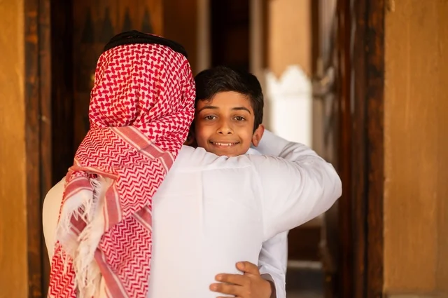 Hugging and greeting at the door is a concept of love and longing, a concept of fatherhood and caring for children. A family stands at the entrance of the house, a Saudi Arabian Gulf father wearing traditional attire and a headscarf looks at his son, a child dressed in white, family bonding.