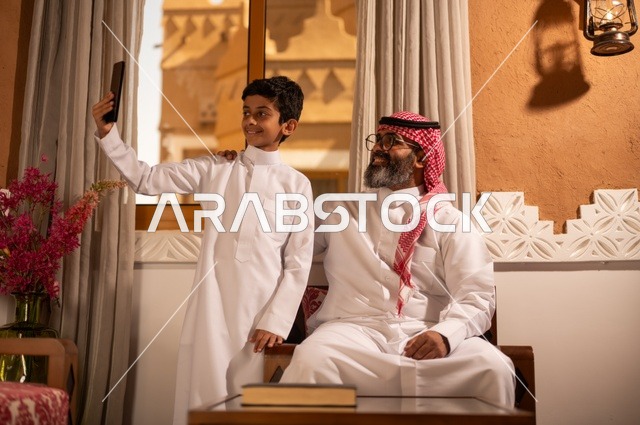 أسرة سعيدة، سيلفي تذكاري للحظة مليئة بالحب والحنان، مفهوم الأبوة والاعتناء بالأبناء, عائلة تجلس داخل مجلس تراثي بديكورات وزخارف تقليدية، أب عربي خليجي سعودي يرتدي الثوب التقليدي والغتره ينظر إلى الكاميرا مع ولده