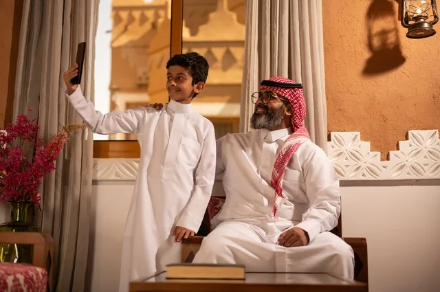 أسرة سعيدة، سيلفي تذكاري للحظة مليئة بالحب والحنان، مفهوم الأبوة والاعتناء بالأبناء, عائلة تجلس داخل مجلس تراثي بديكورات وزخارف تقليدية، أب عربي خليجي سعودي يرتدي الثوب التقليدي والغتره ينظر إلى الكاميرا مع ولده