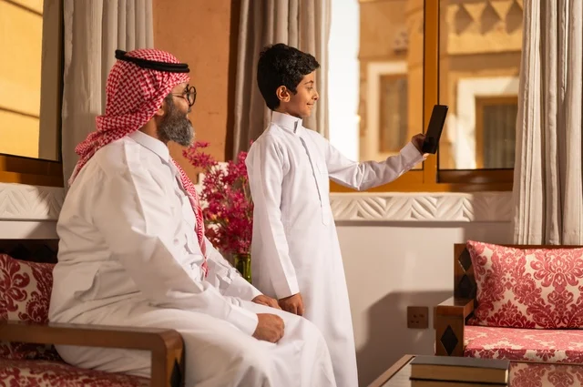 A commemorative selfie of a moment filled with love and tenderness, the concept of fatherhood and caring for children, a family sitting inside a traditional council with traditional decorations and ornaments, an Arab Gulf Saudi father wearing traditional attire and a headscarf looking at the camera with his son, a happy family.