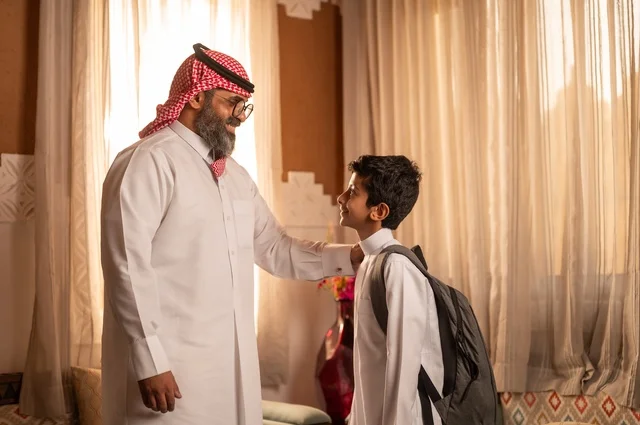 Happy family moments, the concept of fatherhood and caring for children, a family standing inside a traditional council with traditional decorations and ornaments, an Arab Gulf Saudi father wearing traditional attire and a headscarf, placing his hand on his son's shoulder, looks filled with love and tenderness, strolling and spending time with family.