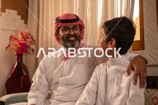 مفهوم الأبوة والاعتناء بالأبناء, عائلة تجلس داخل مجلس تراثي بديكورات وزخارف تقليدية، أب عربي خليجي سعودي يرتدي الثوب التقليدي والغتره وينظر ولده ويقدم له النصائح المفيدة، نظرات مليئة بالحب والحنان، التنزه وقضاء الوقت مع العائلة، لحظات عائلية سعيدة.