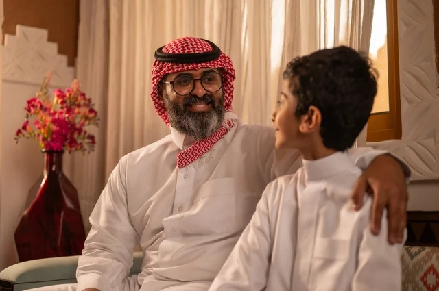 The concept of fatherhood and caring for children, a family sitting inside a traditional council with traditional decorations and ornaments, an Arab Gulf Saudi father wearing traditional attire and a headscarf looking at his son and giving him useful advice, gazes filled with love and tenderness, strolling and spending time with family, happy family moments.