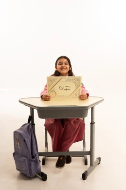 End of the school year, concept of excellence, success and graduation from school, looking at the camera with gestures of pride and happiness, quality of education in Saudi Arabia, portrait of a smiling Saudi Arabian Gulf student wearing school uniform sitting at her desk holding her certificate, white background