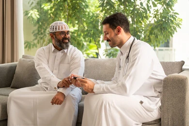 جهاز قياس سكر الدم لمرضى السكري، العمل في القطاع الطبي وشركات التمريض المنزلي، الرعاية الصحية والفحص الطبي السريع, ممرض يرتدي المعطف الطبي يقوم بفحص نسبة السكر في الدم لمريض مسن عربي خليجي سعودي يرتدي الشماغ والثوب التقليدي يجلس على الكنبه