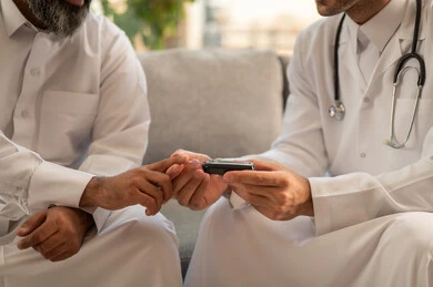 جهاز قياس سكر الدم لمرضى السكري، العمل في القطاع الطبي وشركات التمريض المنزلي، الرعاية الصحية والفحص الطبي السريع, ممرض يرتدي المعطف الطبي يقوم بفحص نسبة السكر في الدم لمريض مسن عربي خليجي سعودي يرتدي الشماغ والثوب التقليدي يجلس على الكنبه