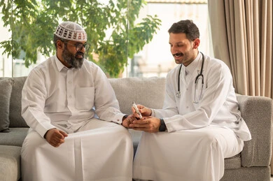 جهاز قياس سكر الدم لمرضى السكري، العمل في القطاع الطبي وشركات التمريض المنزلي، الرعاية الصحية والفحص الطبي السريع, ممرض يرتدي المعطف الطبي يقوم بفحص نسبة السكر في الدم لمريض مسن عربي خليجي سعودي يرتدي الشماغ والثوب التقليدي يجلس على الكنبه