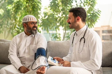 الخدمات الطبية والرعاية الصحية، ممرض يرتدي اليونيفورم والقفازات والكمامة الوقائية يقوم بقياس ضغط الدم لرجل مسن عربي خليجي سعودي يلبس الثوب التقليدي يجلس في المنزل، الإصابة بوعكة صحية، العمل في القطاع الصحي