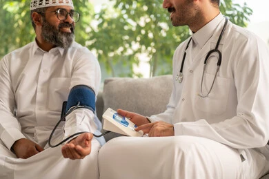 Medical and healthcare services, a nurse wearing a uniform, gloves and a protective mask measuring the blood pressure of an elderly Saudi Arabian Gulf man wearing traditional thobe sitting at home, suffering from a health condition, working in the healthcare sector
