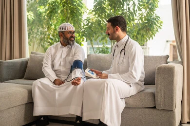 Medical and healthcare services, a nurse wearing a uniform, gloves and a protective mask measuring the blood pressure of an elderly Saudi Arabian Gulf man wearing traditional thobe sitting at home, suffering from a health condition, working in the healthcare sector